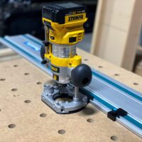 DeWalt trim router mounted on blue Makita guide rail using black and gray plunge base adapter on workbench with pegboard