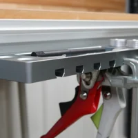 Gray utility rack mounted on aluminum t-track, holding red quick clamps, metal bench dogs, and a black tool in the tray
