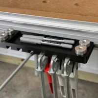 Black utility rack mounted on Festool MFT table track, holding markers and pencils in top tray, with four quick clamps and bench dogs in side slots