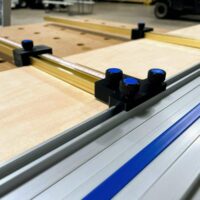 Black plastic parallel guide bracket with three blue knurled knobs mounted on aluminum Kreg ACS guide rail, positioned over plywood workpiece