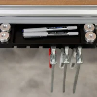 Black utility rack mounted to aluminum t-track, holding two pens and three bench dogs in metal slots, with threaded posts at each corner