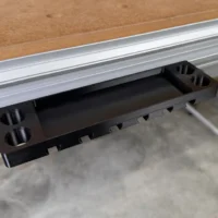 Black plastic utility rack mounted on aluminum t-track, with slots for clamps along the bottom and cylindrical holes on each end
