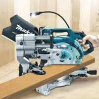 Makita LS610DZ miter saw held in hand while cutting a wooden board, showing the compact blue and silver design with dust collection bag and LED work light