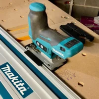 Makita 12v jig saw with turquoise guide rail adapter mounted on a Makita track rail, positioned on a perforated work surface