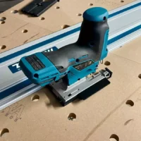 Makita 12v jig saw attached to an aluminum track rail on a perforated work table, showing the blue adapter mounted on the saw's base