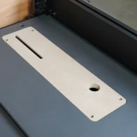Light wood MDF zero clearance insert with centered blade slot and round hole, mounted on dark table saw bed with mounting screws visible