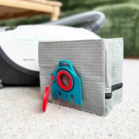 Macam reusable dust bag for Miele vacuums in light gray fabric with blue and red clip attachment, displayed next to a Miele vacuum canister