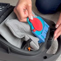 Macam reusable dust bag in blue and red, held in hand above an open Miele vacuum canister with gray fabric interior