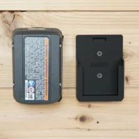 Black ToolCurve battery mount holder next to a Ridgid 18v battery on wood surface, showing the slim profile and two screw holes for wall mounting