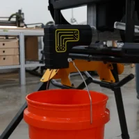 Black plastic adapter with yellow logo mounted on DeWalt table saw, directing sawdust into red bucket suspended below by wire loops