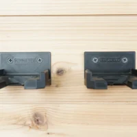 Two dark gray battery mounts for Ryobi 18v batteries mounted on light wood, showing screw holes and clip mechanisms that grip batteries from the sides