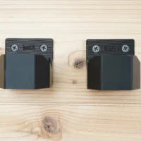 Two black battery mounts with M12 markings and Phillips screw holes mounted on light wood, showing side-profile view