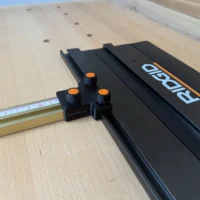 Black parallel guide adapter with orange knobs attached to Ridgid track saw rail, positioned next to tape measure on perforated workbench