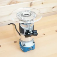 Clear 6-inch round acrylic router base plate mounted on a blue Hercules trim router standing on a workbench