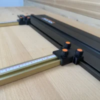 Black plastic parallel guide bracket with three orange knurled knobs mounted on brass-colored Incra T-Track rail, positioned perpendicular to a Ridgid track saw
