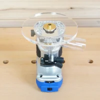 6-inch round clear acrylic router base plate mounted on blue Kobalt trim router, with brass center bushing and black mounting hardware visible