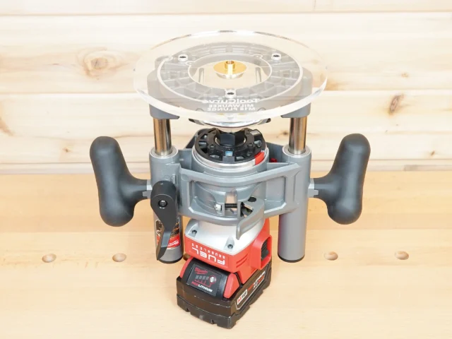 8-inch clear acrylic router base plate mounted on Milwaukee M18 2-1/4HP router, showing circular top with center bit opening and brass insert, viewed from above on workbench