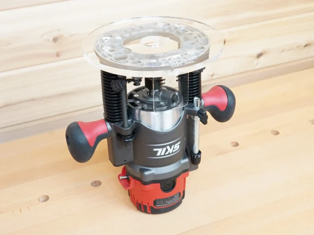8-inch clear acrylic router base plate mounted on a red and black Skil plunge router, showing the enlarged round platform and dual handles on a wooden workbench