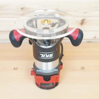 8″ round acrylic router base plate mounted on a Skil RT1322 plunge router, clear from top with brass center fitting and guide bushing holes visible