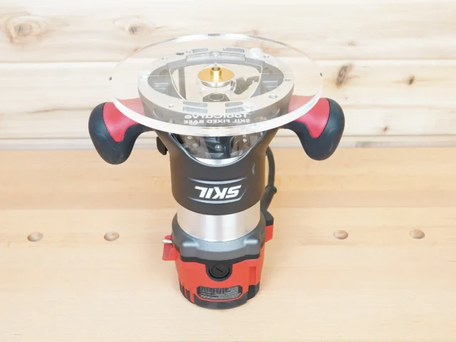 8″ round acrylic router base plate mounted on a Skil RT1322 plunge router, clear from top with brass center fitting and guide bushing holes visible