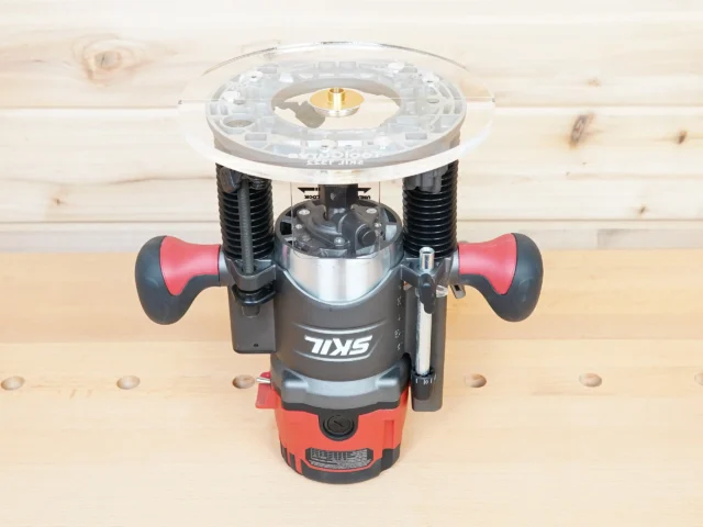 Clear acrylic router base plate mounted on a Skil RT1322 plunge router, showing the 8-inch round base from above with brass threaded insert at center